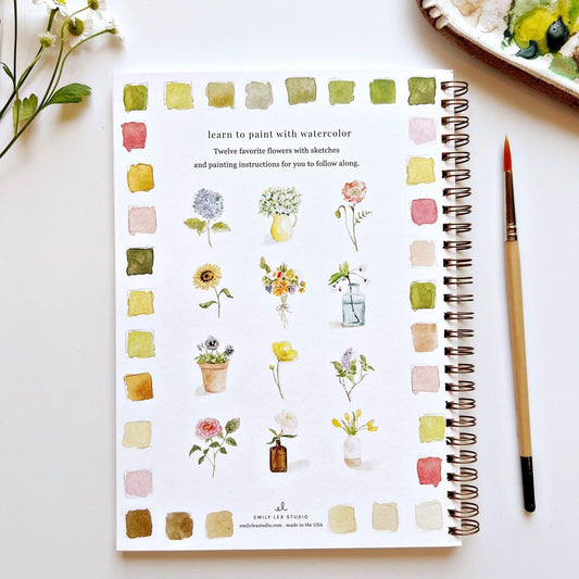 Watercolor painting workbook with watercolor flower illustrations and a paintbrush on a white surface