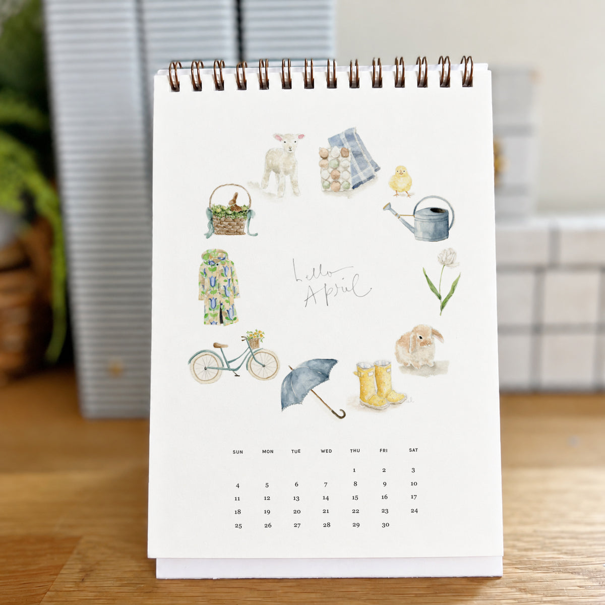 2027 hello month desk calendar April page with watercolor spring icons like a lamb, bicycle, umbrella, and rain boots
