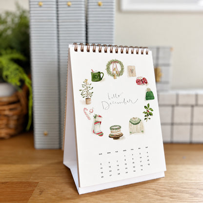 2027 hello month desk calendar December page with watercolor Christmas icons like a wreath, stocking, and sweater