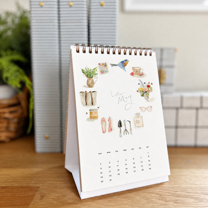 2027 hello month desk calendar May page with watercolor icons like a bird, teacup, ballet flats, and garden tools