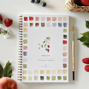 A spiral-bound watercolor workbook titled 'FRUIT' by Emily Lex Studio, displayed on a white surface with fruit props, green leaves, blueberries, a paintbrush, and a woven basket.